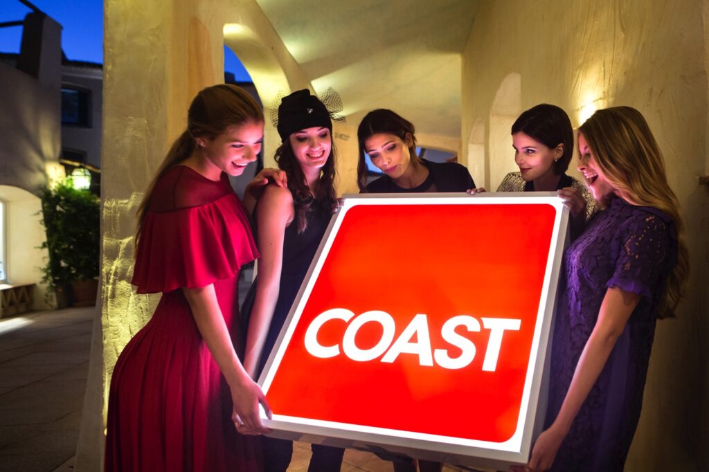 modelle attorno ad una insegna luminosa con il logo della rivista Coast